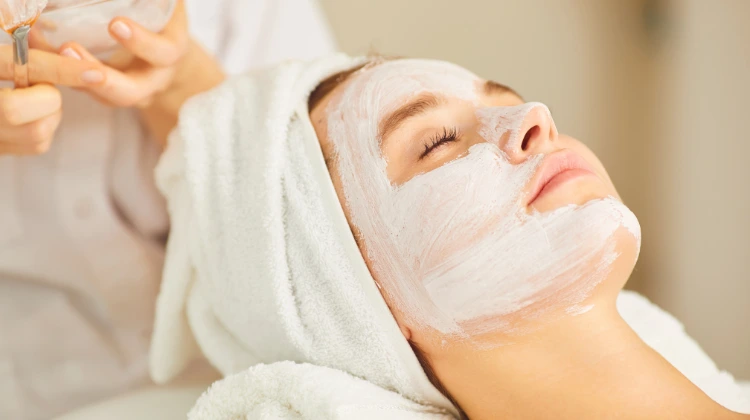 A girl with a towel wrapped around their head is lying down with a white facial mask applied to her face during an antioxidant therapy skincare treatment.