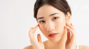 Woman with light makeup gently touches her face, looking thoughtful. Her hair is neatly tied back, happy after having hydrafacial